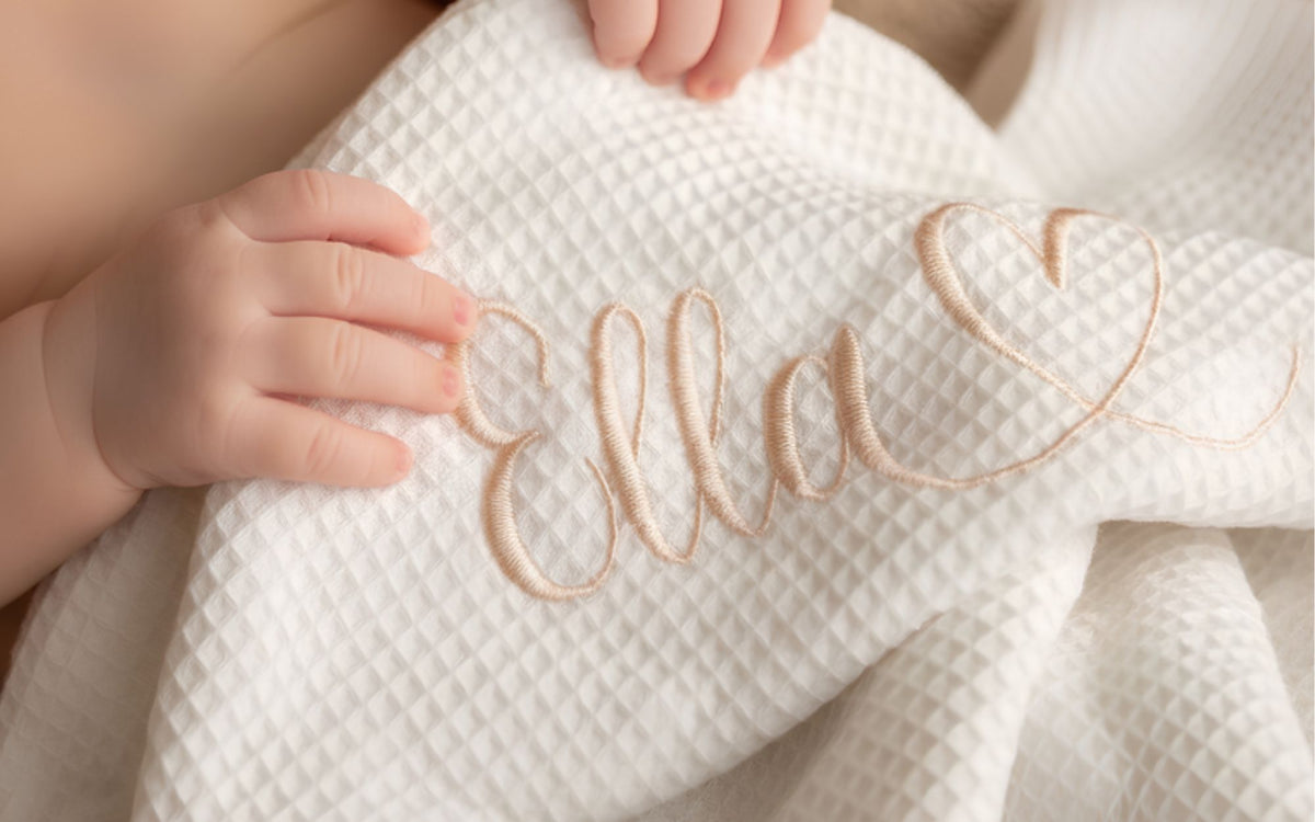 Personalisierte Babydecke aus Waffelpiqué mit gesticktem Namen, Babyhand hält die Decke